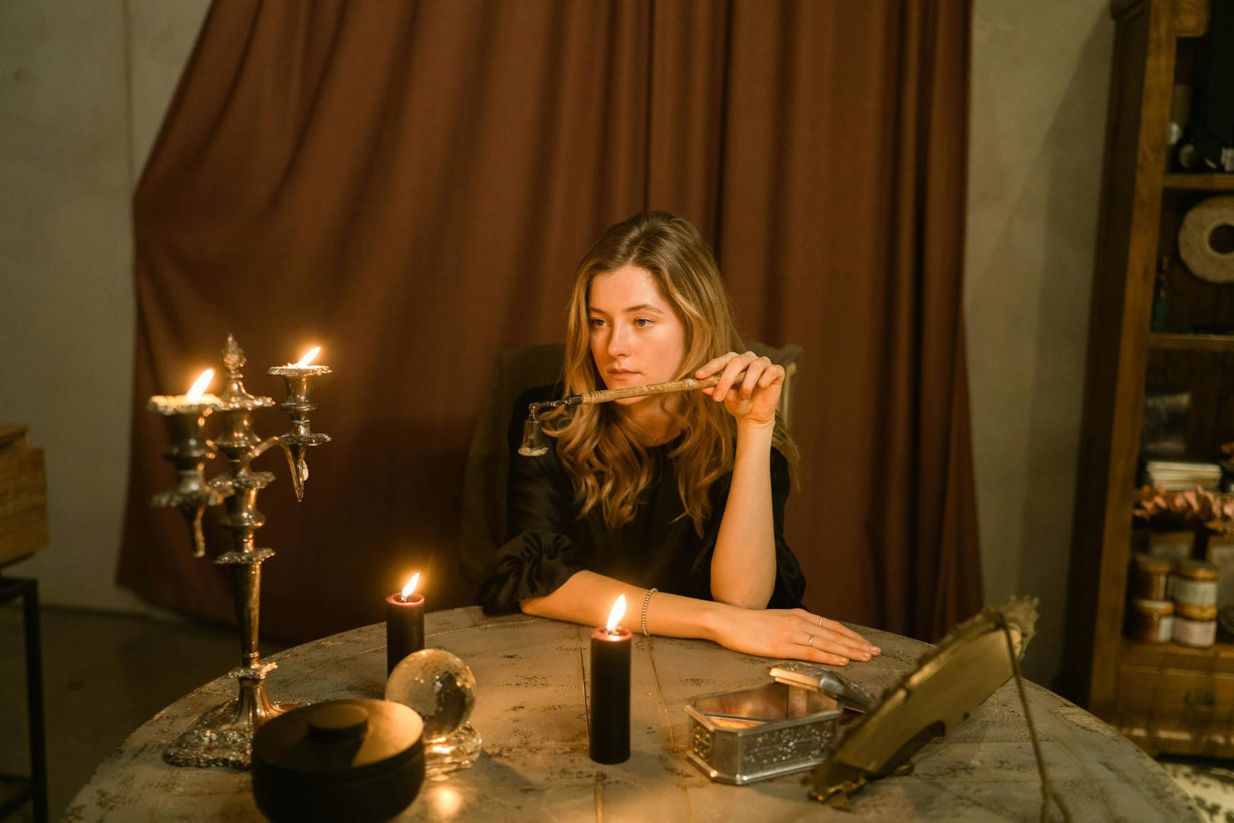 A thoughtful woman sits at a table with candles, engaged in a ritualistic setting, indoors. on The Manifesting Light